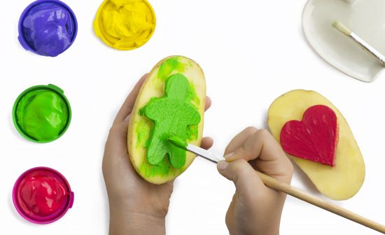 Childs hand, play painting with potato stamps, brushing green on a figure shape, red ,yellow, and purple poster paints  some isolated objects on a white background, copy space