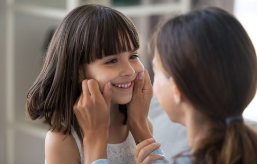 Close up focus on smiling girl, mother touching cheeks of lovely daughter with tenderness and love family spending time indoors. Elder sister adores her small sister, people playing together at home