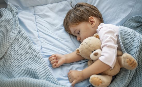 Healthy child, sweetest blonde toddler boy sleeping in bed holding her teddy bear. Adorable toddler girl taking a nap in a grey bed holding her teddy bear. Baby toddler asleep with teddy bear