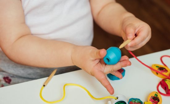 Child putting beads on a string. Bead stringing activity. Fine motor skills development. Early education, Montessori Method. Cognitive skills, children development. Close up of baby's hands.