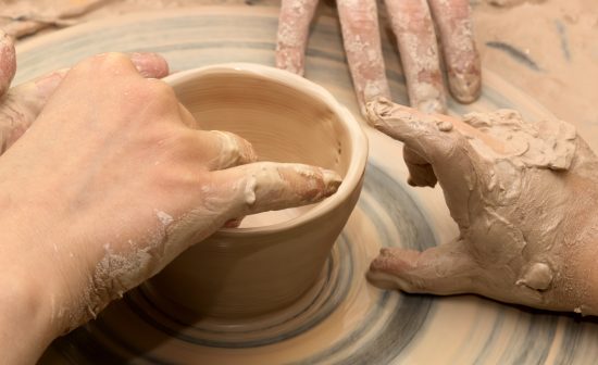 Beginner and teacher hands in clay at process of making crockery on pottery wheel. Potters at work. Panoramic view.