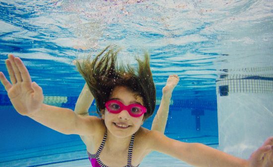 Girl underwater in swimming pool wearing goggles looking at camera smiling